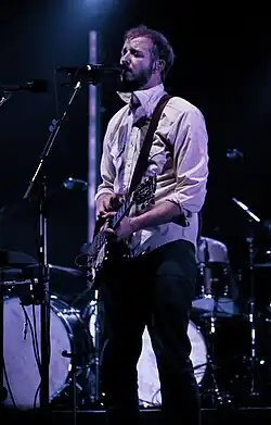 Justin Vernon playing a guitar and singing onto a mic, in dim stage lights