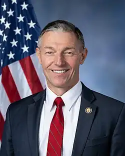 Official House portrait of Kennedy smiling in front of the U.S. flag, wearing a black suit with American flag lapel pin, white shirt, and red tie.