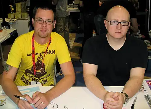 Two men who are cartoonists in a subcultural convention. The man on the left is Mike Krahulik, who draws the comic. He wears glasses and a yellow T-shirt, and has stubbles. On the other hand, another man on the right is Jerry Holkins, who writes the comic. He is a bald man with glasses and a black T-shirt.