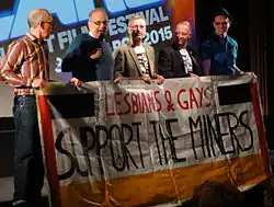5 white men (4 have glasses) stand holding a banner with handwritten text: "LESBIANS & GAYS SUPPORT THE MINERS". The second man is talking on a microphone. In the background a film screen is partially visible with the text "FILM FESTIVAL [...] 2025".
