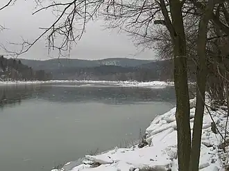 A grey lake under grey skies with snow and ice blocks on its shore and snow covered low mountains in the background