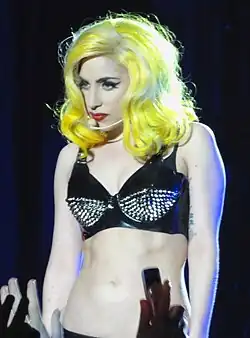 Lady Gaga performing on stage, wearing a black latex bra with spiked embellishments. Her hair is styled in voluminous waves, dyed bright yellow at the ends. She has dramatic winged eyeliner and red lipstick, and she wears a headset microphone while looking intensely at the audience.