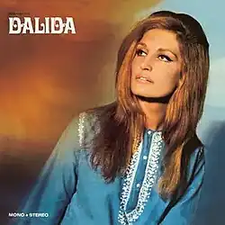 Dalida posing in studio in front of orange background, looking aside she wears 1960s style shirt and her signature minimalistic makeup around eyes.