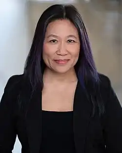 A professional headshot of an Asian American woman in her late 40s, looking into the camera with a neutral smile. She wears a black top and natural-looking makeup, and her long, loose hair has subtle purple highlights.