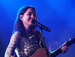 In blue lighting especially visible on her hair, a woman performs onstage with an acoustic guitar and a big smile.