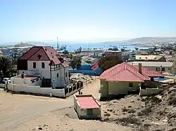 Colour photograph of Lüderitzof taken close to Diamantberg Street. Red tiled houses can be seen in the foreground, and in the background, the Atlantic Ocean can be glimpsed in a bay that is surrounded on sides by rocky land.