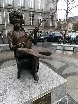 Statue of Luke Kelly sitting on a chair and playing an instrument and (presumably singing). The statue is placed on a cement platform. The platform is on a pavement with benches to sit on. In the background, there is a big brown building, with cars and bicycles parked in front, along with tree (without leaves). There are other smaller buildings on the top right corner away from focus.