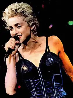 Madonna performing on stage, wearing a dark blue corset with cone-shaped bust cups and black straps. Her short, curly blonde hair is styled in a voluminous look, and she holds a microphone while singing.