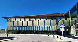 A long, low building glad in vertical panels of glass and metal. The roof line is angular and coppery, with painted swirling designs on the right side. The building is connected by a skywalk bridge to another building out of frame. The text on the front reads Manitou a bi Bii daziigae