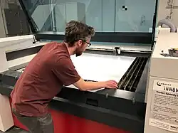 A technologist inserts a sheet of acrylic into a commercial grade laser cutter as part of the manufacturing process to create face shields