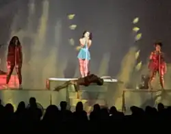 A color photograph of Marina performing live with backup singers and singing into a microphone.