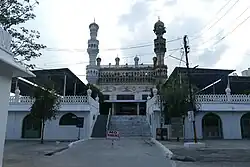 A richly decorated mosque is seen situated on top of a platform, with a fight of steps leads to it. The mosque is flanked by two minarets. One of the minarets is whitewashed, while the rest of the mosque is cream-colored. There are two white buildings, with arched entrances and windows on the ground level, on either side of the flight of steps. Two small trees are also seen in the foreground.