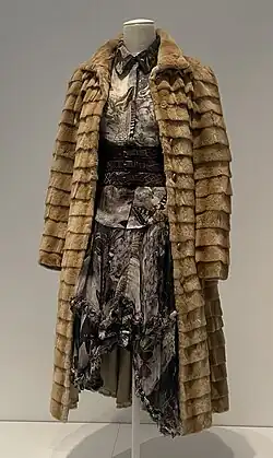 A light brown fur coat worn over a blouse and skirt in a fabric printed with birds, moths, and skulls