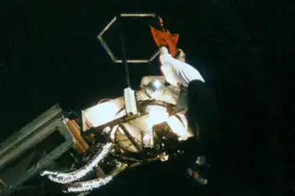 A person with pale skin dangling by their hands from a crane. The person is wearing a white shirt and black pants and footwear. A black background can be seen behind the person.