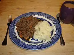 A blue patterned plate atop a table and flanked by a knife and fork, containing a portion of mince and tatties served with a cup of tea