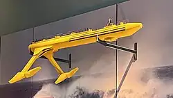 Looking up at a model of the yellow SR2000 tidal turbine mounted on brackets from ta wall. The turbine has a long tubular hull, with two angled legs protruding down from the end, each of which has a nacelle and two-bladed rotor
