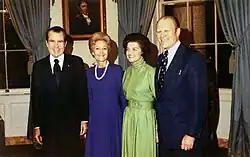 Two women are flanked by two men in suits, standing in a room of the White House.