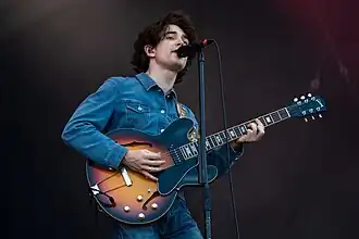 A poorly-shaved pale man with thick black curly hair with a microphone stand to his mouth wearing a jean jacket and holding a black and brown acoustic guitar standing in front of a black background.