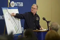 A man with glasses wearing a blue shirt standing at a lectern with a microphone points toward a map on an easel to his left.