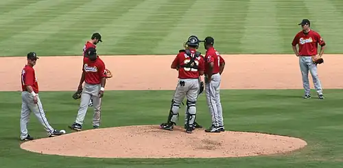 Seven men wearing red baseball jerseys, gray pants, and black caps are standing on a baseball diamond's infield; three are talking together on the mound.