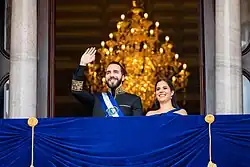 A photograph of Nayib Bukele and Gabriela Rodríguez waving from a balcony