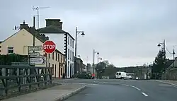 A picture of the Main Street in Newbliss, featuring the junction between the R183 and R189. Road signs for Monaghan, Three Mile House, and Aghabog are visible, along with a stop sign.