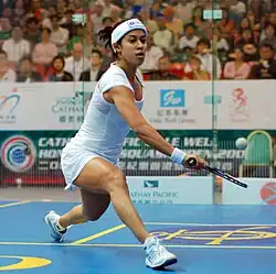 A young female squash player in white, stretching out to get her racket under a low ball, on a blue court with yellow markings, in front of a large, out-of-focus crowd behind glass.