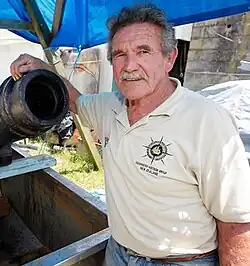 Hilliam, a burly man with a silver moustache, leaning on the muzzle of a cannon underneath a tarpaulin shelter.