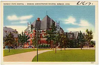 Image ID, a vintage illustrated postcard with a tan border. The top left inscription reads “Norwich State Hospital – Showing Administration Building, Norwich, Conn.” In the top right corner is the number 2. Between the two is handwritten, “6/6/7.” The Administration Building is a bright red, likely brick structure. It is in the French Neo-Gothic style. It has three stories, with two white lines between each. The roof is heavily sloped with a spike at each of its three peaks. There is a chimney protruding from the shortest of the roofs. To the left of the building are five parked cars and another brick building. Behind the Administration Building is another similar building. In front of it is a lawn with an arched tan path lined with trees and street lamps. The sky is a calm blue with a few clouds.