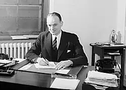 Black and white photograph of John Dickey in a suit seated at a desk, writing on papers, with office furniture and a window in the background.
