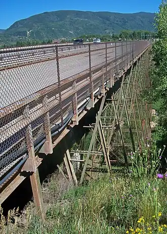 An old metal bridge across a grassy, wooded gorge, seen from its right. Diagonal supports go down below the bridge. There is a wooded mountain in the background.