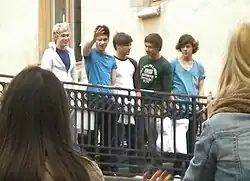 Five teenage boys greeting and waving at fans from a balcony. From left to right: Niall Horan, Zayn Malik, Louis Tomlinson, Liam Payne, and Harry Styles.