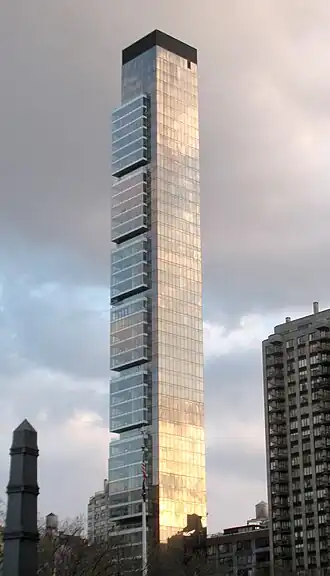 A tall, thin building with some slight squarish projections at its higher levels seen from between some trees.