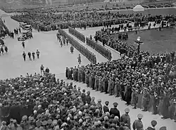 Black and white photo: Crowds of people and a ranked military guard gather to watch four men in ceremonial uniform approach a set of stone steps.