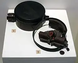 Black disc-shaped landmine on a white display stand, alongside broken fragments of another landmine