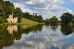 A colour photograph of a lake with a ruined Gothic building standing on the bank on the left and a vineyard on the sloping hillside behind