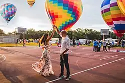Man twirls a woman in a long dress in front of hot air balloons lifting off during sunrise.