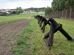 View along a row of triangular metal spikes standing between a bare earth strip and a metal fence