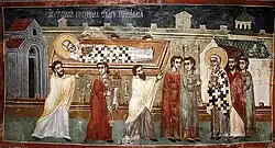 Painting of three white men carrying a coffin with a white haloed bearded man in it. Three head tops are visible beyond the coffin. They are preceded by two white tonsured men in robes holding sticks (candles? lances?). They are received by a haloed white bearded man and two acolytes. Cyrillic text is over the coffin.