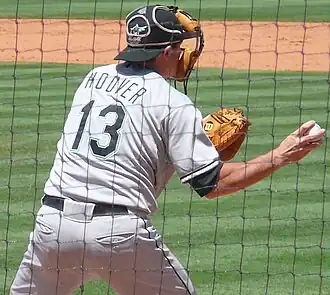 A man in a gray baseball uniform reading "Hoover" and "13" on the back and a black catcher's mask on his face throwing a baseball with his right hand