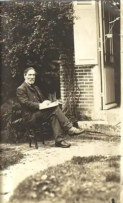 Photograph of Paul Milliet, older than during the events of the Commune. He is sitting on a chair outside a house, holding a book in his hands.