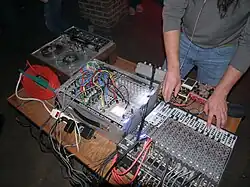 A photo of Pete Swanson (only lower part of his body is visible) performing live, with various pieces of electronic equipment placed on a table in front of him.