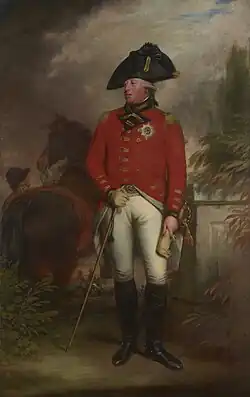 Clean-shaven man wearing the red jacket of an 1800 British army general with the star of the Order of the Garter, white breeches, black knee-high boots, and a black bicorne hat. Behind him a groom holds a horse.