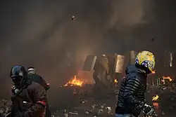 Three helmeted protestors throwing pavement bricks at riot-police line under concealment of smoke from burning tires