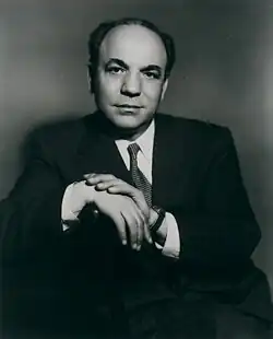 A man in a dark suit with a checkered tie sits on a chair sideways, hands at rest atop the chair's back.