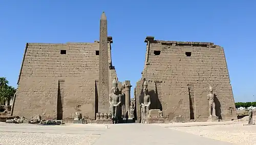 Pair of trapezoidal stone towers flanking a passage, beyond which a row of columns is visible. In front of the towers are several large statues and an obelisk.