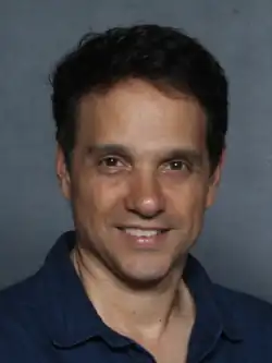 A dark-haired man wearing a blue shirt poses for the camera in a blank background