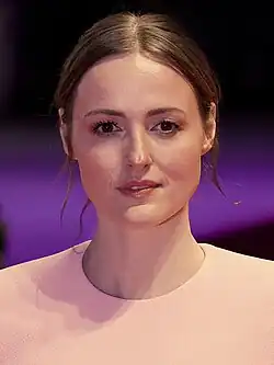 A woman with brown hair wearing light clothing in front of a purple background
