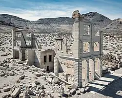 Aerial view of the same three-story white masonry bank building in 2022, roofless with large sections of its walls missing. Masonry rubble lies about the building, which has openings for doors and windows but no glass or wood. The collapsed walls of another, smaller ruin are to the right. Barren hills are visible in the distance.