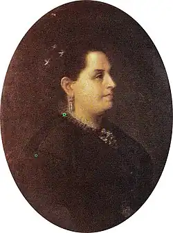 Portrait of a middle-aged woman with tan skin, dark eyes, and dark hair dressed in an updo with jeweled ornaments at the crown, she is wearing a dark dress, drop earrings, and an necklace